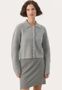 Svetlošedý rebrovaný kardigán s golierom a šiestimi gombíkmi, doplnený čierno-bielou sukňou v vzor houndstooth. Úzky strih, jemná textúra.