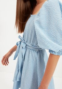 Modrá a bílá kostkovaná šaty s nafouknutými rukávy, čtvercovým výstřihem a látkovým páskem uvázaným v pase. Měkká bavlněná textura.