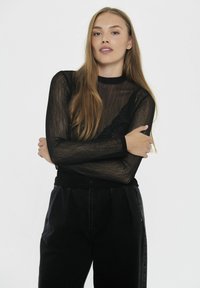 Top negro transparente de manga larga con un acabado texturizado y un escote ajustado, combinado con pantalones negros de cintura alta. La modelo posa con los brazos cruzados.