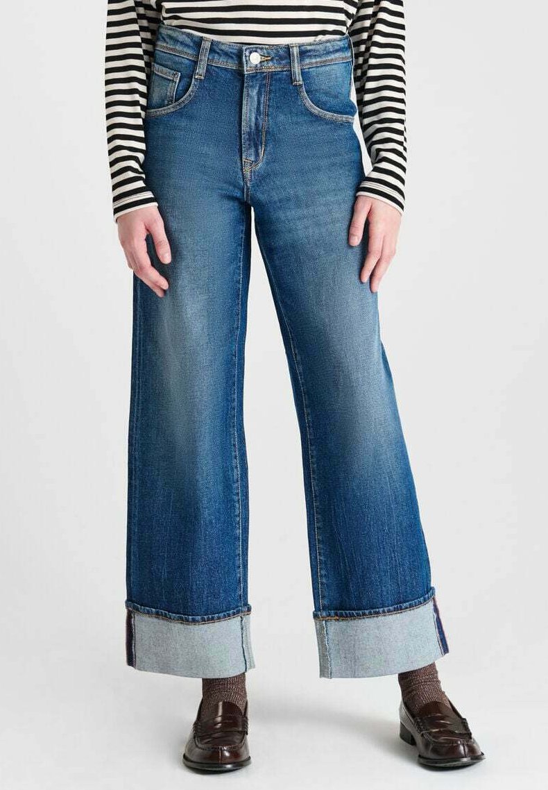 Jean en denim bleu à jambes larges et taille haute, avec des ourlets contrastants, associés à des mocassins bruns et un haut rayé.