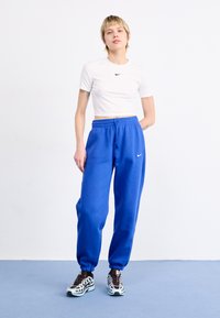 Weiße, kurze T-Shirt mit schwarzem Nike-Logo, kombiniert mit blauen Sweatpants mit elastischem Bund und Bündchen sowie auffälligen Sneakers.