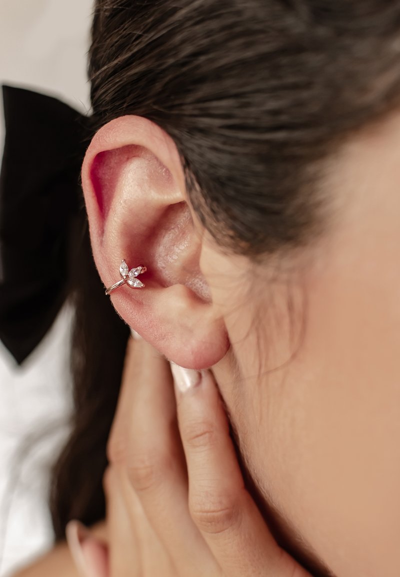 Pendiente tipo ear cuff en oro rosa con un diseño floral y piedras claras en corte marquesa, presentado en un primer plano de una oreja. Texturas suaves evidentes.