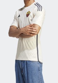 Camisa de fútbol color crema con un patrón sutil, con cuello redondo, acentos laterales grises y el logo de AS Roma en el pecho.
