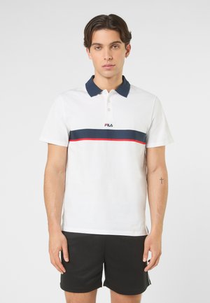 Giovane uomo con capelli corti e scuri indossa una polo bianca con striscia blu navy e rossa e pantaloncini neri, in piedi davanti a uno sfondo semplice.