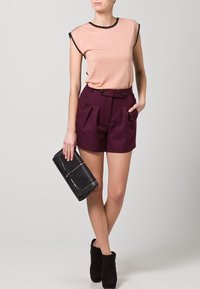 Peach short-sleeve top with black trim, paired with high-waisted maroon shorts featuring double buttons and pleats, accessorized with a black clutch.
