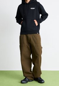 Sweat à capuche noir avec logo blanc, associé à un pantalon cargo marron ample. Des chaussures de sport noires complètent la tenue.