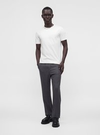 Homme debout portant un t-shirt blanc à manches courtes, un pantalon gris, des chaussures noires et un collier chaîne argenté sur un fond clair uni.