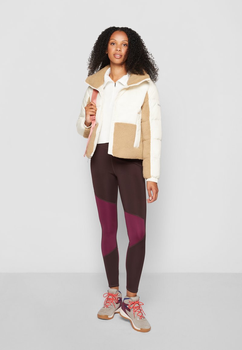 Giubbotto puffer in crema e beige, con una grande tasca frontale; abbinato a leggings marrone scuro con dettagli color bordeaux e sneakers chiare.