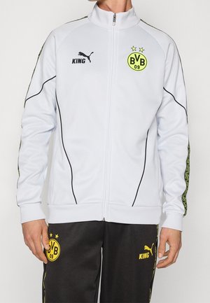 Homme portant une veste de sport blanche zippée et un pantalon noir avec les logos jaunes BVB 09 et Puma King.