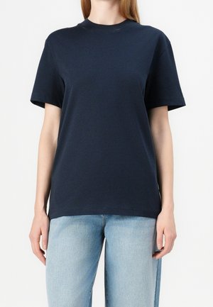 Femme portant un t-shirt uni bleu marine foncé à manches courtes et col rond, et un jean bleu clair sur fond blanc.