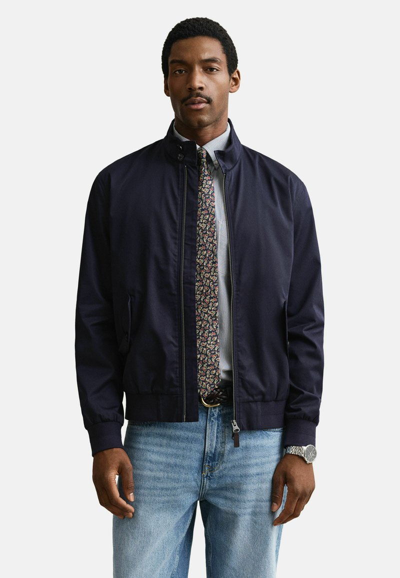 Homme portant une veste bomber bleu marine sur une cravate à motifs et une chemise rayée, avec un jean bleu clair et une montre-bracelet au poignet gauche.