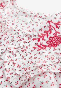 Weiße Bluse mit floralen Motiven und kleinen roten Blumen, Rüschendetails und roten bestickten Akzenten. Sie verfügt über eine Knopfblende mit einem "NEXT"-Label.
