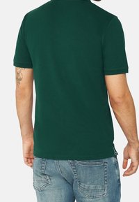 Tmavě zelené polo tričko s krátkým rukávem, rovný lem, boční rozparky a měkkou bavlněnou texturu. Obsahuje jednoduchý límec a žádné viditelné logo.