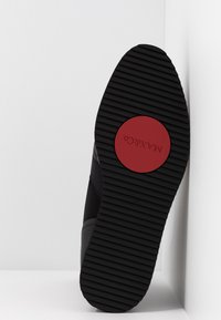 Semelle de chaussure noire avec des rainures horizontales et un logo circulaire rouge marqué "MAX&Co" au centre, positionnée sur un fond blanc.