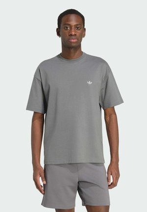 Homme portant un t-shirt gris Adidas et un short assorti, debout devant un fond clair uni, regardant devant avec une expression neutre.