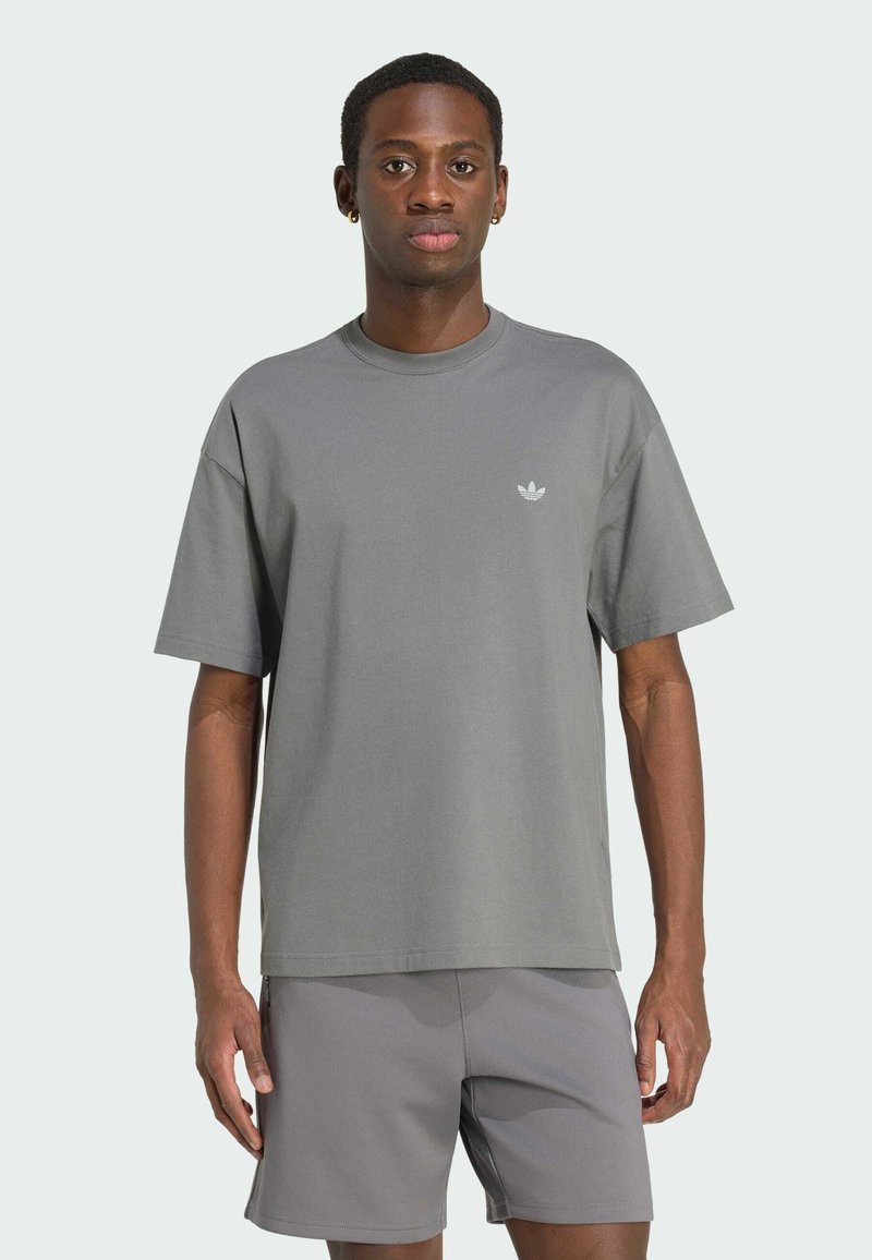Homme portant un t-shirt gris Adidas et un short assorti, debout devant un fond clair uni, regardant devant avec une expression neutre.