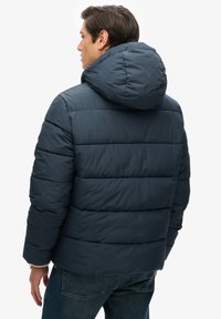 Mørkeblå dunjakke med hætte, med horisontale quiltede sektioner og en glat tekstur. Båret over denimjeans.