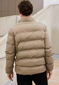 Geacă puffer bej cu panouri matlasate orizontale, guler înalt și manșete elastice. Textură moale și netedă, concepută pentru căldură.