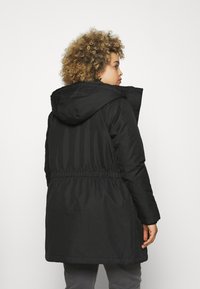 Manteau noir rembourré avec capuche, taille cintrée et manches longues. Présente une texture lisse et un design long jusqu'aux genoux.