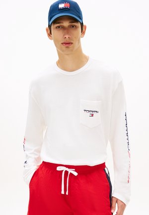 Giovane uomo con cappellino Tommy Jeans blu navy, maglia bianca a maniche lunghe con logo e pantaloncini rossi con coulisse, su sfondo bianco uniforme.