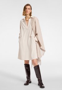 Beiges Kleid mit Kragen, tiefem V-Ausschnitt und Taschen an der Seite, kombiniert mit einem hellbeigen Schal mit Fransen und getragen mit glänzenden braunen Stiefeln bis zum Knie.