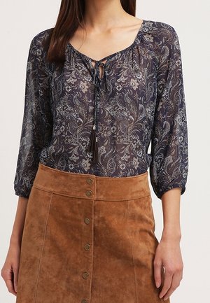 Žena nosí tmavou halenku se vzorem paisley s tříčtvrtečními rukávy a hnědou semišovou sukni na knoflíky.