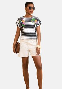 Striped short-sleeve shirt in navy and white with floral embroidery, paired with white shorts and a brown handbag. Oversized sunglasses present.