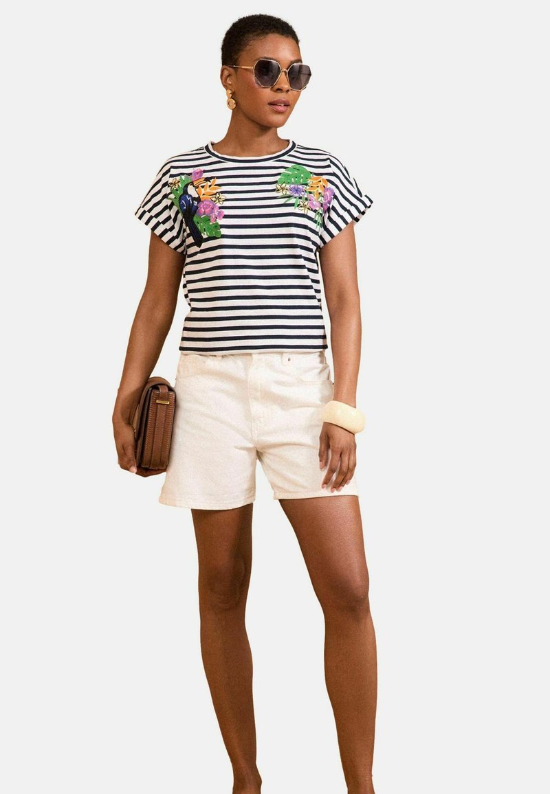 Striped short-sleeve shirt in navy and white with floral embroidery, paired with white shorts and a brown handbag. Oversized sunglasses present.