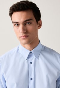 Ljusblå skjorta med knappslå och subtilt texturerat mönster, med spetsig krage och mörkblå knappar. Passar för formella eller lediga tillfällen.