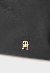 Sac à main en nylon noir à texture lisse, avec un logo en métal doré. Design rectangulaire simple sans motifs supplémentaires.