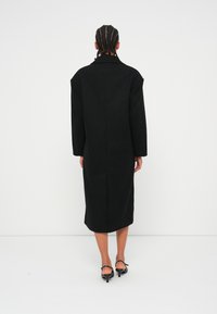 Manteau long noir avec une silhouette structurée, des épaules larges et un col cranté. Il présente une couture au dos et est fabriqué dans un tissu lisse.