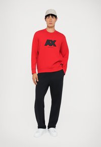 Červená mikina s čiernym logom, skombinovaná s čiernymi teplákmi a bielymi teniskami. Model má na hlave svetlosivú čiapku. Jednoduchý, ležérny outfit.