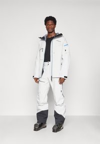 Chaqueta impermeable blanca con detalles negros, cremallera y bolsillos; combinada con pantalones blancos con secciones inferiores grises y botas de esquí negras.
