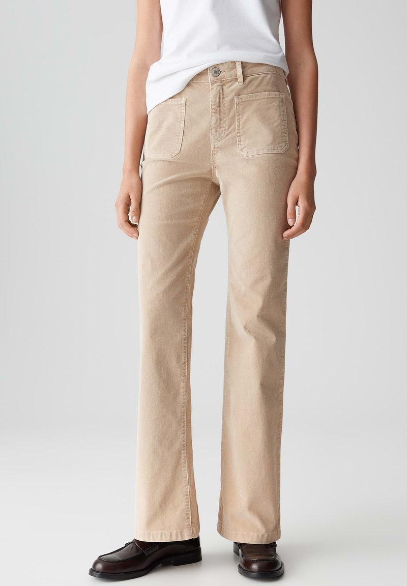 Persona che indossa pantaloni beige a gamba dritta con grandi tasche anteriori, una maglietta bianca e mocassini marroni scuri, in piedi contro uno sfondo chiaro e semplice.