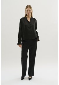 Schwarze Seidenbluse mit lockerer Passform, Knopfleiste und gefältelten Ärmelabschlüssen, kombiniert mit schwarzen taillierten Hosen und spitz zulaufenden High Heels.