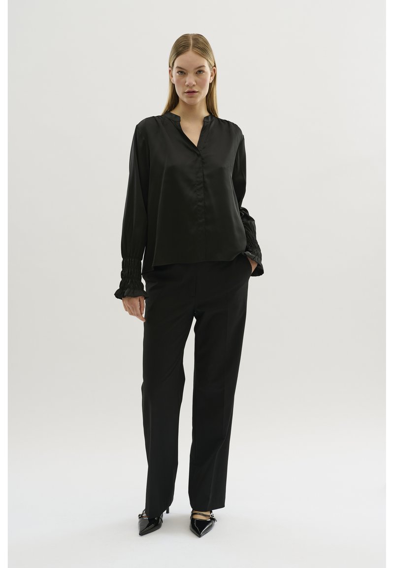 Schwarze Seidenbluse mit lockerer Passform, Knopfleiste und gefältelten Ärmelabschlüssen, kombiniert mit schwarzen taillierten Hosen und spitz zulaufenden High Heels.