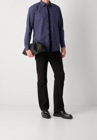 Marinblå långärmad skjorta, knäppt, med spetsig krage, kombinerad med svarta raka jeans och svarta skor. Håller en rullad svart väska.