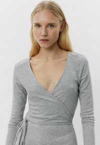 Grauer langärmliger Wickeloberteil aus gerippter Textur mit tiefem V-Ausschnitt und seitlichem Bindedetail. Es hat eine körperbetonte Silhouette und einen glatten Stoff.