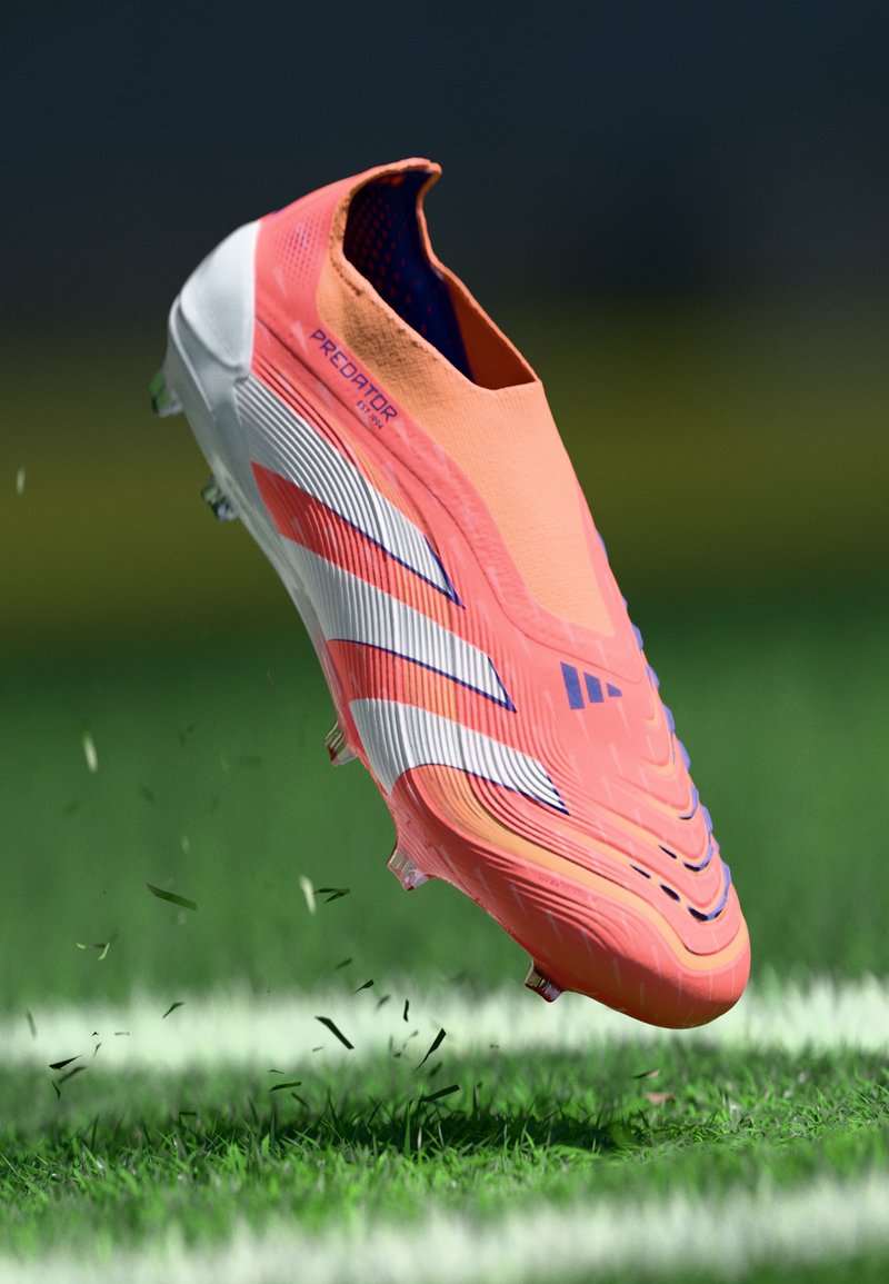 Scarpa da calcio arancione brillante con strisce bianche, tomaia testurizzata e tacchetti a forma di cono. Design leggero, sospesa sopra un prato verde.