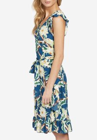 Kvinna iklädd en blå blommig omlottklänning med volangärmar och volang i nederkant, knuten vid midjan, stående i profil mot en vit bakgrund.