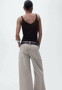 Canottiera marrone con spalline sottili, abbinata a jeans beige a gamba larga con una cintura nera. Tessuto morbido con dettagli sulle tasche posteriori.