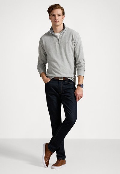 Grå zip-up genser, hvit skjorte under, mørke jeans, brune joggesko og brunt belte. Modellen står mot en nøytral bakgrunn.