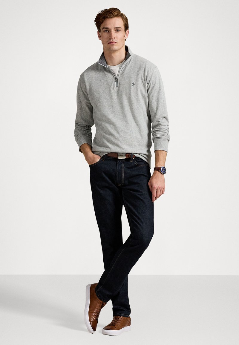 Suéter gris con cremallera, camisa blanca debajo, jeans oscuros, zapatillas marrones y un cinturón marrón. El modelo está de pie contra un fondo neutral.