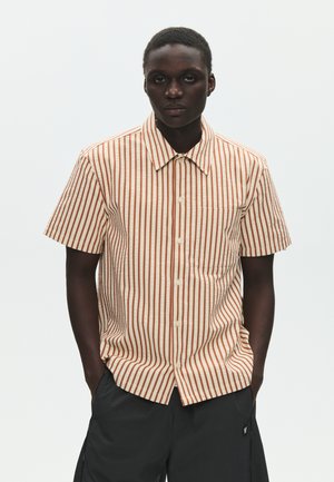 Hombre con camisa de manga corta abotonada a rayas verticales beige y naranja y pantalones negros, de pie con las manos en los bolsillos frente a un fondo liso.