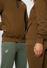 Bruine sweatshirt met capuchon en zwarte trekkoorden; groene joggingbroek met logo. Zachte stof, geribbelde boorden en taillebanddetails.