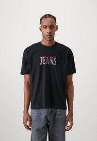 Camiseta negra de algodón con la palabra "guess" en rojo cursiva y "JEANS" en gris, cuello redondo, mangas cortas, con un ajuste relajado, combinada con jeans grises.