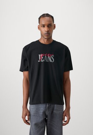 Juoda medvilnės marškinėliai su raudonai užrašytu "guess" ir pilkai "JEANS", suapvalintu kaklu, trumpomis rankovėmis ir laisvu siluetu, derinami su pilkais džinsais.