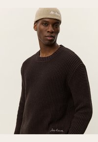 Dark brown textured knit sweater with a round neckline, paired with a light beige knitted beanie featuring white embroidered text.