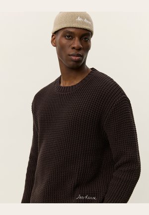 Dark brown textured knit sweater with a round neckline, paired with a light beige knitted beanie featuring white embroidered text.