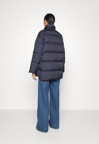 Marineblaues Puffer-Jacke mit horizontalem Steppmuster, hohem Kragen und oversized Passform, kombiniert mit weit geschnittenen blauen Jeans.